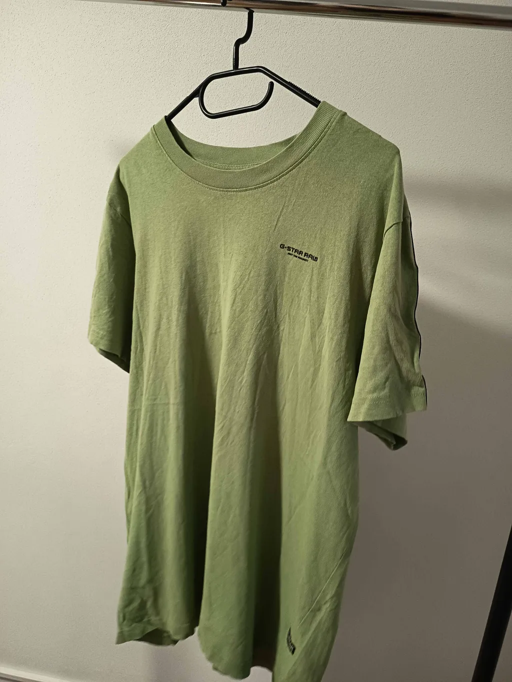 Phone photo of a green T-shirt on a hanger, shot at an angle against a white wall